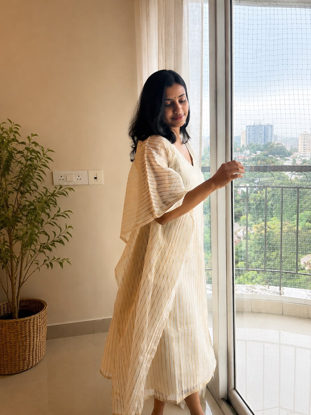 Ponnila Kaftan in ivory and gold stripe, worn by a window